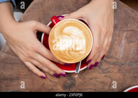 Gros plan de mains féminines tenant un café expresso sur une table. Le café est décoré d'art latte complexe et servi dans une tasse rouge. Vue de dessus Banque D'Images