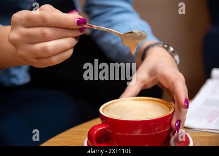 Gros plan d'une main de femme tenant une petite cuillère avec de la mousse d'espresso épaisse en cascade dans sa tasse de café rouge sur une table de café Banque D'Images