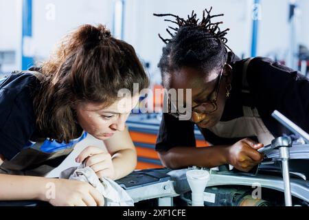 Mécaniciens afro-américains collaborant à l'entretien du véhicule cassé, en vérifiant si le radiateur est défectueux. Femmes multiethniques dans l'atelier de réparation automobile travaillant ensemble sur la réparation de voiture, discutant des meilleures options Banque D'Images