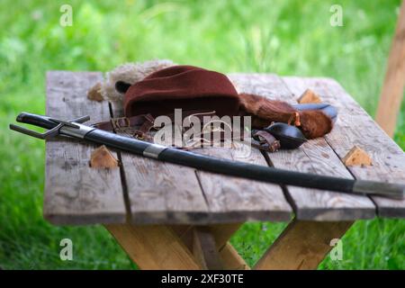 Équipement de reconstitution médiévale sur une table en bois. Les articles comprennent une épée dans un fourreau noir, un chapeau de laine marron, un bracelet en cuir et un accessoire en fourrure, Set ag Banque D'Images