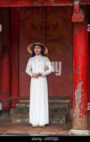 Jolie jeune femme vietnamienne portant une robe traditionnelle Ao Dai et un chapeau conique debout dans une porte ornée au Temple de la littérature, Hanoi, Nord Vietnam Banque D'Images