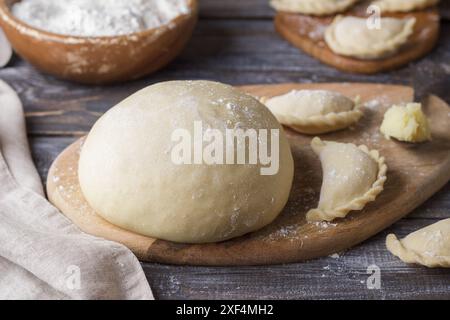 Pâte pour boulettes sur une planche à découper en bois, boulettes prêtes à l'emploi, un bol de farine et une cuillère avec remplissage de pommes de terre sur une table en bois Banque D'Images