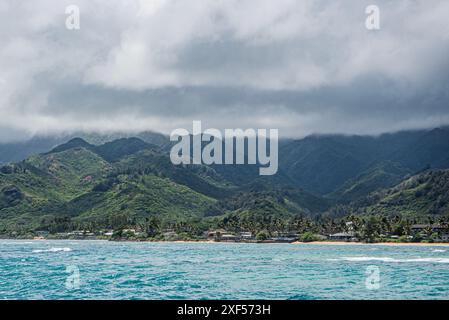 Une vue impressionnante sur le paysage hawaïen avec une mer bleu clair, une plage de sable blanc et d'imposantes montagnes bordées de palmiers. Cette scène encapsulat Banque D'Images