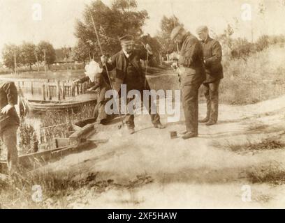 Allemagne circa.1900 – Un groupe d'hommes et une femme, debout sur une rive de rivière, pêchant. Ils utilisent des cannes à pêche traditionnelles en bois. La femme porte un grand bonnet blanc en coton. Trois des hommes placent des appâts sur leurs lignes de pêche. Banque D'Images