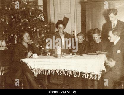 Allemagne. 1920s. Une famille s'est réunie autour d'une table en regardant à travers les albums de photos au moment de Noël. Debout derrière est un grand arbre de Noël décoré. Une théière ornée sur un support avec un brûleur est sur la table, avec une bouteille de liqueur en grès et des verres à boire. Banque D'Images