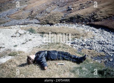 Suisse. 1960 – Une jeune femme portant un pantalon tartan, une veste de moto et des bottes, dort au soleil près d’un ruisseau, avec une carte pliée à côté. Banque D'Images