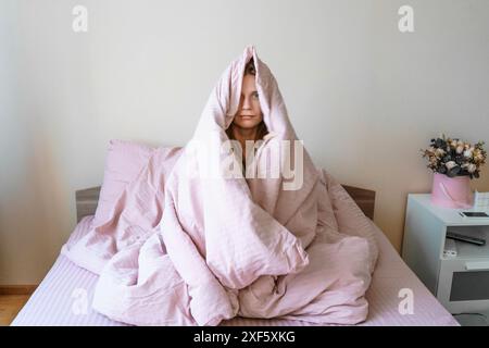 la femme est assise sur le lit avec une couverture rose la recouvrant. regarder la caméra avec une expression sérieuse Banque D'Images