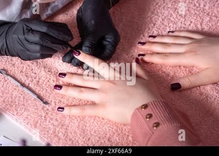 Esthéticienne peignant les ongles de ses clients avec un gel spécial de paillettes cyclamen dans un salon de beauté. La table est recouverte d'une serviette. Concept de beauté Banque D'Images