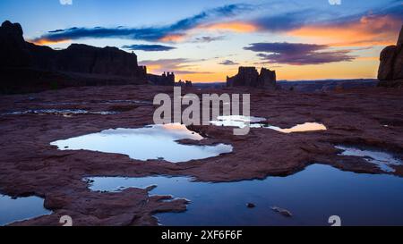 La nuit tombe au parc national des Arches avec des flaques d'eau de pluie dans les rochers au crépuscule tandis que les nuages brillent de couleurs chaudes Banque D'Images