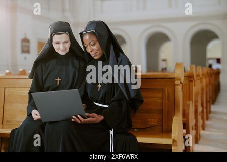 Plan moyen de deux religieuses jeunes et matures ethniquement diverses assis sur un banc en bois dans une église catholique travaillant sur un ordinateur portable Banque D'Images