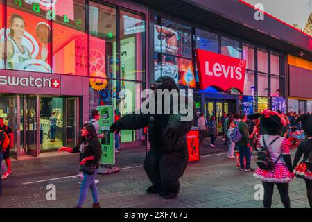 Vue sur la rue animée de Times Square, avec des gens vêtus de costumes, y compris des costumes de gorille, à l'extérieur des magasins Swatch et Levi's avec des lumières vives Banque D'Images