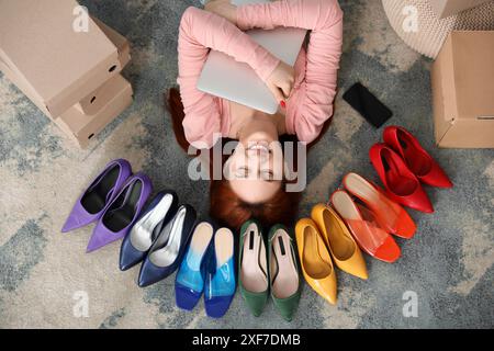 Jeune femme couchée sur le sol avec ordinateur portable et téléphone portable parmi les talons hauts dans la chambre Banque D'Images