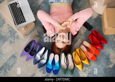 Jeune femme allongée sur le sol avec ordinateur portable, téléphone portable et faire du cœur avec des mains entre des talons hauts dans la chambre Banque D'Images