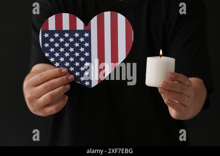 Femme tenant la bougie brûlante et le drapeau des États-Unis en forme de coeur sur fond sombre, gros plan. Journée nationale de prière et de souvenir pour les victimes de Banque D'Images