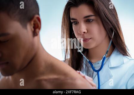 Un jeune médecin ausculte les poumons d'un patient masculin lors d'un examen médical. Le médecin porte un manteau blanc et a un stéthoscope autour Banque D'Images