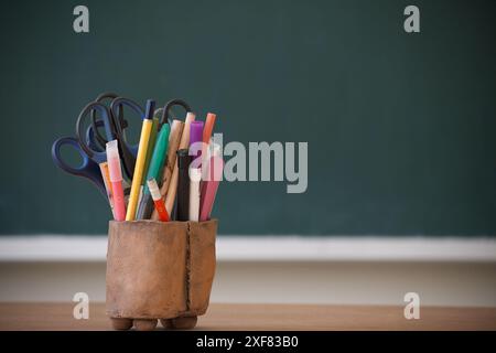 Une variété de fournitures pour la salle de classe, y compris des stylos, des marqueurs et des ciseaux, organisés dans un support en bois devant un tableau noir. Banque D'Images