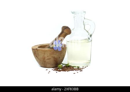Mortier en bois rempli de petites graines de lin brun et une plante de lin bleu fleurit à côté de l'huile de lin dans une carafe en verre isolée sur fond blanc Banque D'Images