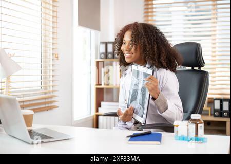 Un médecin féminin noir professionnel et confiant dans une robe blanche a une consultation en ligne avec son patient, montrant la radiographie au patient, WOR Banque D'Images