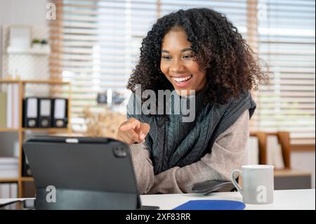 Une femme noire attrayante et joyeuse dans un pull tricoté confortable pointe son doigt vers un écran de tablette tout en ayant une réunion en ligne avec son collègue Banque D'Images