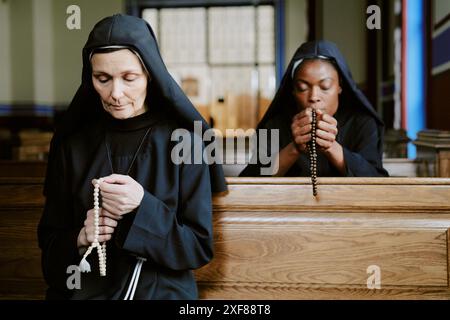 Deux religieuses catholiques jeunes et matures ethniquement diversifiées assises sur des bancs à l'église, priant avec le chapelet dans les mains Banque D'Images