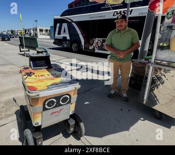 West Hollywood, Californie, États-Unis. 30 juin 2024. Oscar, un robot Axel 'Service Robotics' sans pilote, est vu conduire lui-même rapidement à travers West Hollywood le 30 juin 2024, alors qu'il passe devant un bus avec LA sur le côté, et aussi le mot 'Dash'', devant un stand de fruits coloré avec un mexicain dans une chemise verte amusé par le chariot de robot autonome nommé Oscar. Un panneau en arrière-plan indique « Big Outdoor » et il y a deux coeurs jaunes sur le trottoir. Il s'est arrêté près d'un stand de fruits coloré, sous un panneau d'affichage qui montre un homme prenant une photo et un qui dit, 'Dashâ (crédit image Banque D'Images