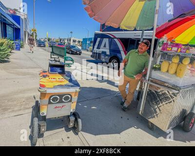 West Hollywood, Californie, États-Unis. 30 juin 2024. Oscar, un robot Axel 'Service Robotics' sans pilote, est vu conduire lui-même rapidement à travers West Hollywood le 30 juin 2024, alors qu'il passe devant un bus avec LA sur le côté, et aussi le mot 'Dash'', devant un stand de fruits coloré avec un mexicain dans une chemise verte amusé par le chariot de robot autonome nommé Oscar. Un panneau en arrière-plan indique « Big Outdoor » et il y a deux coeurs jaunes sur le trottoir. Il s'est arrêté près d'un stand de fruits coloré, sous un panneau d'affichage qui montre un homme prenant une photo et un qui dit, 'Dashâ (crédit image Banque D'Images