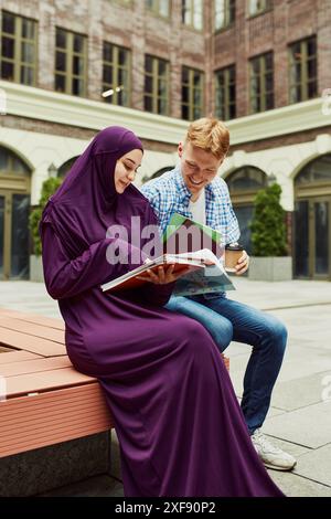 Amis souriants, étudiants, fille musulmane et garçon caucasien assis à l'extérieur du campus et se préparant à la leçon ensemble. Banque D'Images