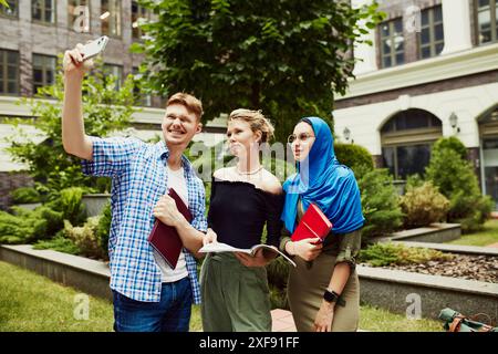 Premier jour d'université, faire des souvenirs. Jeunes, garçons et filles prenant selfie, debout à l'extérieur du campus avec des livres, caucasiens et musulmans Banque D'Images