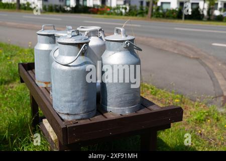 Quatre canettes de lait argentées reposent sur un banc en bois brun foncé, positionné près d'une rue calme. Banque D'Images