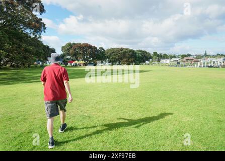 Homme marchant sur la pelouse de Milford Beach Reserve en été. Tentes et canopée pop-up sous les arbres Pohutukawa en fleurs. Auckland. Banque D'Images