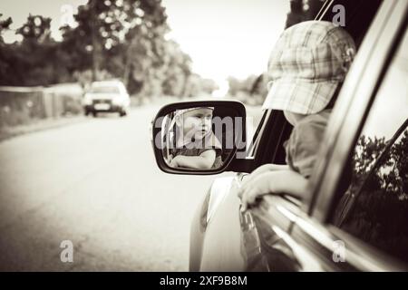 Un enfant portant un chapeau regarde curieusement leur reflet dans le rétroviseur latéral d'une voiture sur une route avec des arbres en arrière-plan. Biélorussie, Minsk Banque D'Images