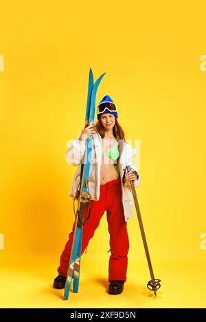 Fille combinant des vêtements d'été et d'hiver, tenant du matériel de ski, posant sur fond de studio jaune vif Banque D'Images