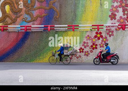 Vendeur de fleurs vendeur de fleurs vélo d'équitation par mur de mosaïque suivi de près par moto cyclomoteur à Hanoi, Ha Noi, Nord Vietnam, Asie en juin Banque D'Images