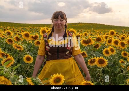 Une femme se tient debout au milieu d'un champ avec beaucoup de tournesols jaunes. Cheveux tressés, portant une blouse jaune, une robe de soleil brune et un tablier jaune chez su Banque D'Images