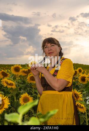 Une femme se tient debout au milieu d'un champ avec beaucoup de tournesols jaunes. Cheveux tressés, vêtus de vêtements nationaux allemands, chemisier jaune, sundr brun Banque D'Images