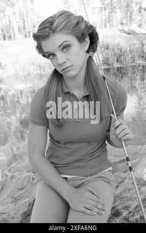 Adolescente cente caucasienne assise sur un rocher près d'un étang, tenant une canne à pêche et bobine, profitant d'une journée paisible de pêche. ÉTATS-UNIS Banque D'Images