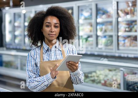 Jeune femme employée debout dans une allée d'épicerie, utilisant une tablette numérique pour la gestion des stocks. Elle porte une tenue décontractée avec un tablier Banque D'Images