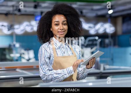 Jeune travailleuse dans le département de poisson d'un magasin utilisant une tablette. Elle porte un tablier et une chemise à carreaux, illustrant le service de vente au détail professionnel dans un cadre moderne. Banque D'Images
