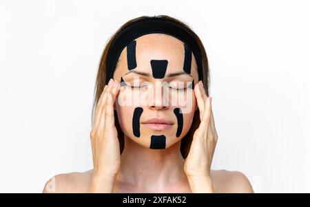 Portrait d’une mannequin de beauté appliquant du ruban kinesio noir sur son visage, mettant en valeur une technique de soin tendance et innovante. Ruban adhésif pour le contour du visage. Banque D'Images