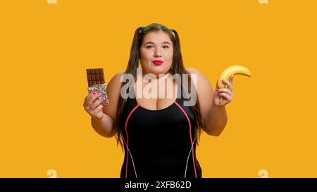 Nourriture de choix. Problème nutritionnel. Sinueuse femme confuse en surpoids doute manger de la banane utile ou du chocolat au lait savoureux malsain posant isolé sur fond jaune. Banque D'Images