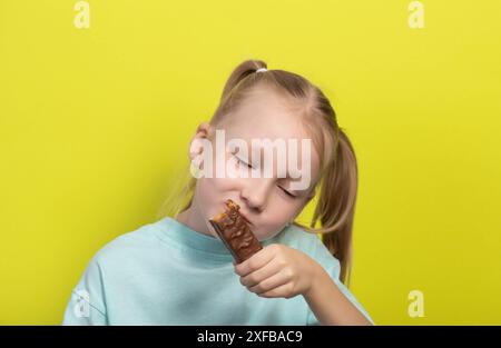 Une belle fille joyeuse aux cheveux blancs de sept ans mange une barre de chocolat sur fond jaune. Les enfants sont des amoureux de la malbouffe sucrée, horizonto Banque D'Images