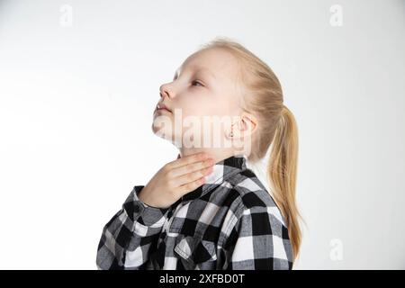 Une jeune fille blonde âgée de sept ans lui tient mal à la gorge avec ses mains. Maux de gorge et allergies chez l'enfant. Inflammation des ganglions lymphatiques, asthme Banque D'Images