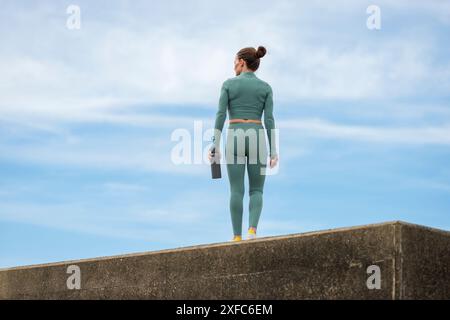 Vue arrière d'une femme sportive tenant une bouteille d'eau, s'entraînant à l'extérieur, fond de ciel bleu. Banque D'Images