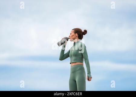 Femme sportive prenant un verre d'eau après l'exercice à l'extérieur, fond de ciel bleu. Banque D'Images
