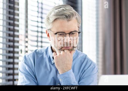 Un homme d'affaires mature en lunettes et une chemise bleue réfléchit profondément tout en travaillant sur un ordinateur portable dans un bureau moderne. L'image transmet la concentration, la réflexion et le dévouement. Banque D'Images