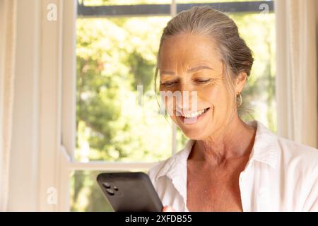 Femme âgée souriante à l'aide d'un smartphone, profitant du temps de loisirs à la maison près de la fenêtre Banque D'Images