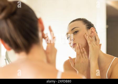 Application de soins de la peau, jeune femme examinant son visage dans le miroir de la salle de bain Banque D'Images