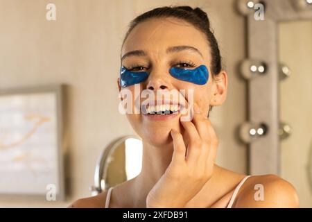 Application de patchs sous les yeux, jeune femme souriant et touchant le visage à la maison Banque D'Images