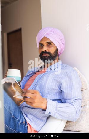 Détente à la maison, homme en turban lisant livre et tenant tasse à café Banque D'Images