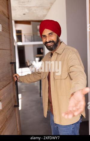 Invité accueillant, homme indien souriant dans turban ouvrant la porte à la maison Banque D'Images